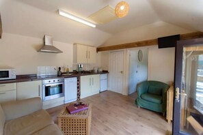 A compact kitchen with white cabinets, a microwave, and a stove. There is a green armchair and a wooden crate with items on it. - Tranquil Cottage in a Countryside (Ashford)