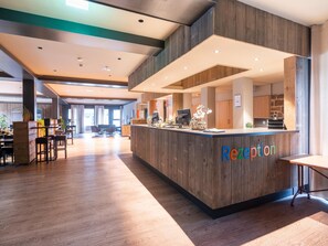 Ein Empfangsschalter mit Holzoptik und der Aufschrift 'reception'. Im Hintergrund sind Tische und Stühle zu sehen, und der Raum ist modern gestaltet, mit Holzböden und einer hohen Decke.
