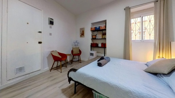 Una camera da letto con un letto, una sedia, una scrivania, una ventola e scaffali.