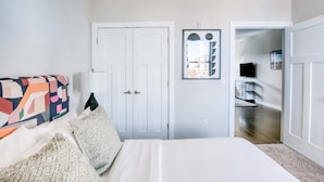 Um quarto com uma cama, um sofá colorido, uma porta branca e um corredor que dá para outro cômodo.