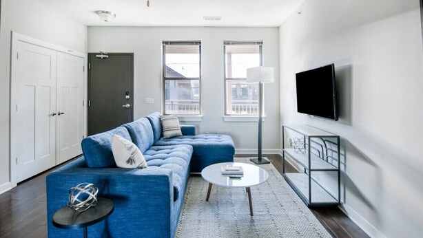 Uma sala de estar moderna com um sofá seccional azul, uma mesa de centro redonda, uma TV de tela plana na parede e uma grande janela que oferece luz natural.
