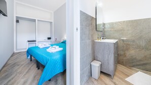 Hotellihuone, jossa on sänky, yöpöytä, pieni työpöytä, peili, kylpyhuone, jossa on pesuallas ja suihku, sekä roskakori.