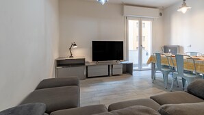 Ein modernes Wohnzimmer mit einem Flachbildfernseher, grauen Sofas und einem Essbereich mit Tisch und Stühlen.