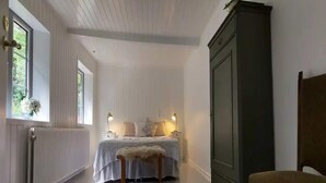 Une chambre à coucher comprenant un lit, des lampes de chevet, un radiateur, une armoire, un banc et une fenêtre donnant sur un paysage verdoyant.