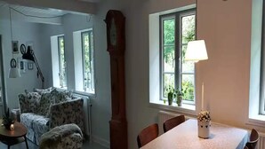 Un salon avec un canapé à motifs floraux, une table à manger avec des chaises, un lampadaire et une horloge comtoise.