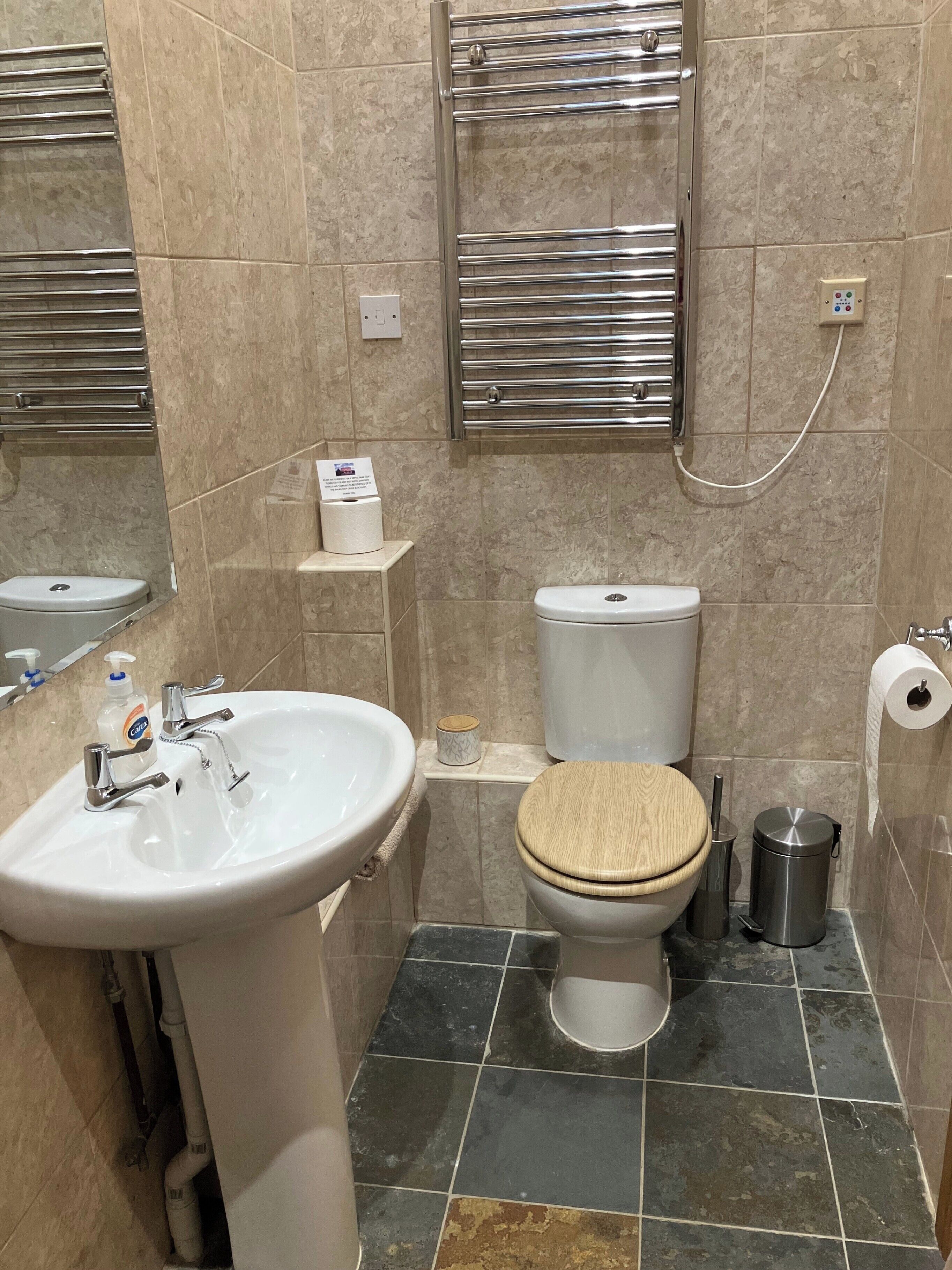 A bathroom with a white sink, a toilet with a wooden seat, a towel rack, and a trash bin.