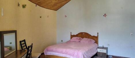 Une chambre avec un plafond en bois, un lit avec une literie rose, une commode en bois, une chaise et un petit tapis.