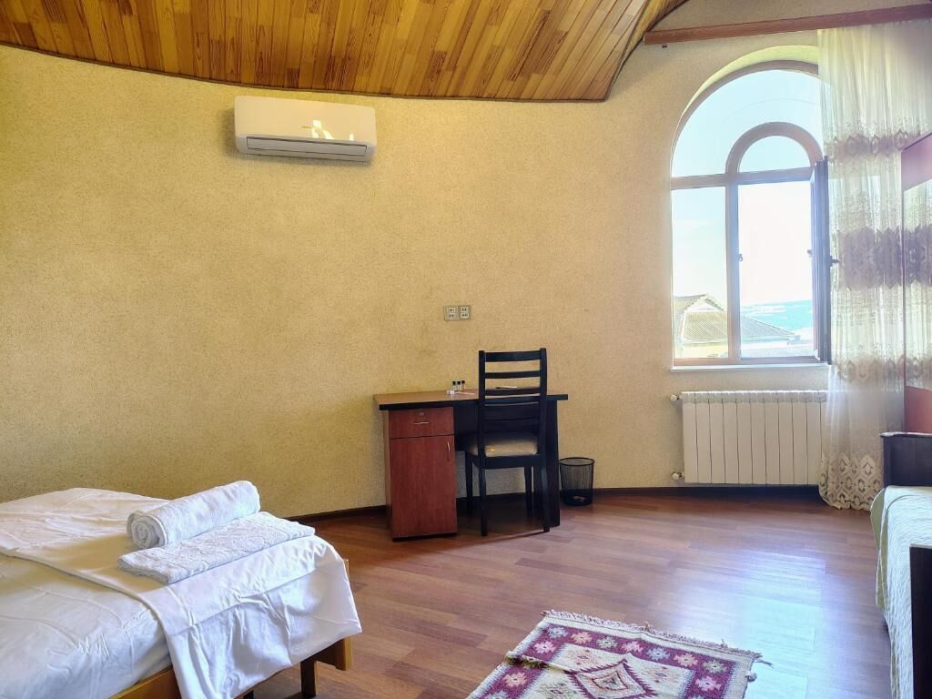 A room with a bed, a desk, and a chair. There is a window with curtains and a view of the outside.