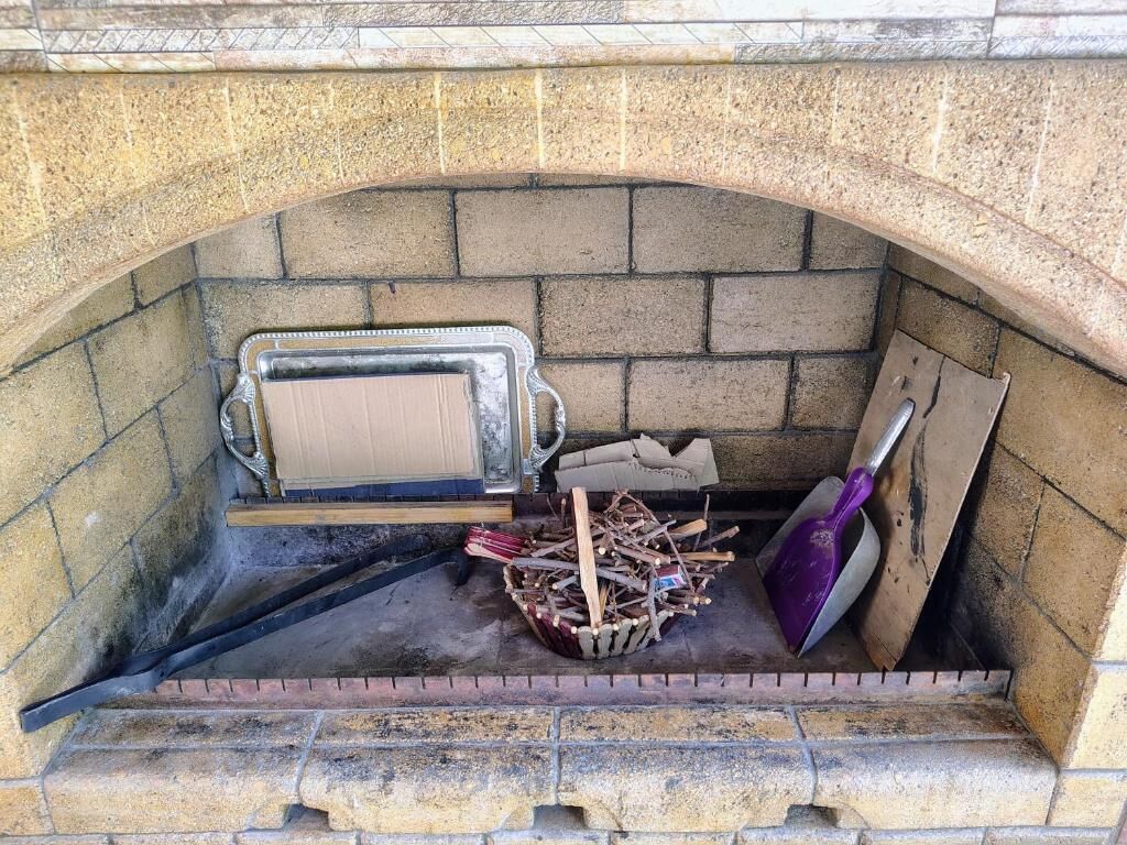A stone-built fireplace with a metal tray, a pile of firewood, and a shovel.