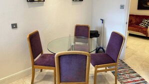 Ein Essbereich mit einem Glastisch, um den vier lilafarbene Stühle stehen. An der Wand hängen zwei gerahmte Kunstwerke und im Hintergrund steht ein rotes Sofa.