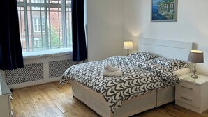Ein Schlafzimmer mit einem Bett, zwei Nachttischen, einem Fenster mit Vorhängen, einem Heizkörper, einem Bild an der Wand und einem Holzboden.