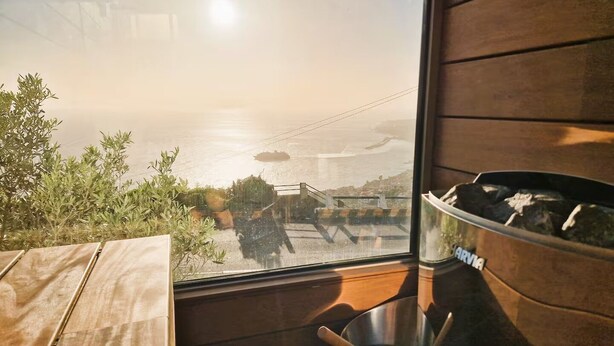 A room with a large window overlooking a coastal area, a wooden table, and a metal bowl. - Shell Living - Infinity Loft (FUNCHAL)