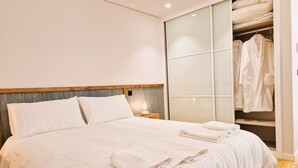 A modern hotel room with a large bed, a wooden headboard, a walk-in closet with robes, and a sliding glass door. - Shell Living - Infinity Loft (FUNCHAL)