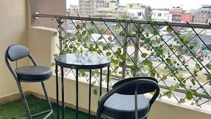 A balcony with artificial grass, two chairs, a small round table, and a view of buildings through a metal fence.