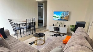 A living room with a sofa set, a dining table with chairs, a TV mounted on the wall, and a small side table.
