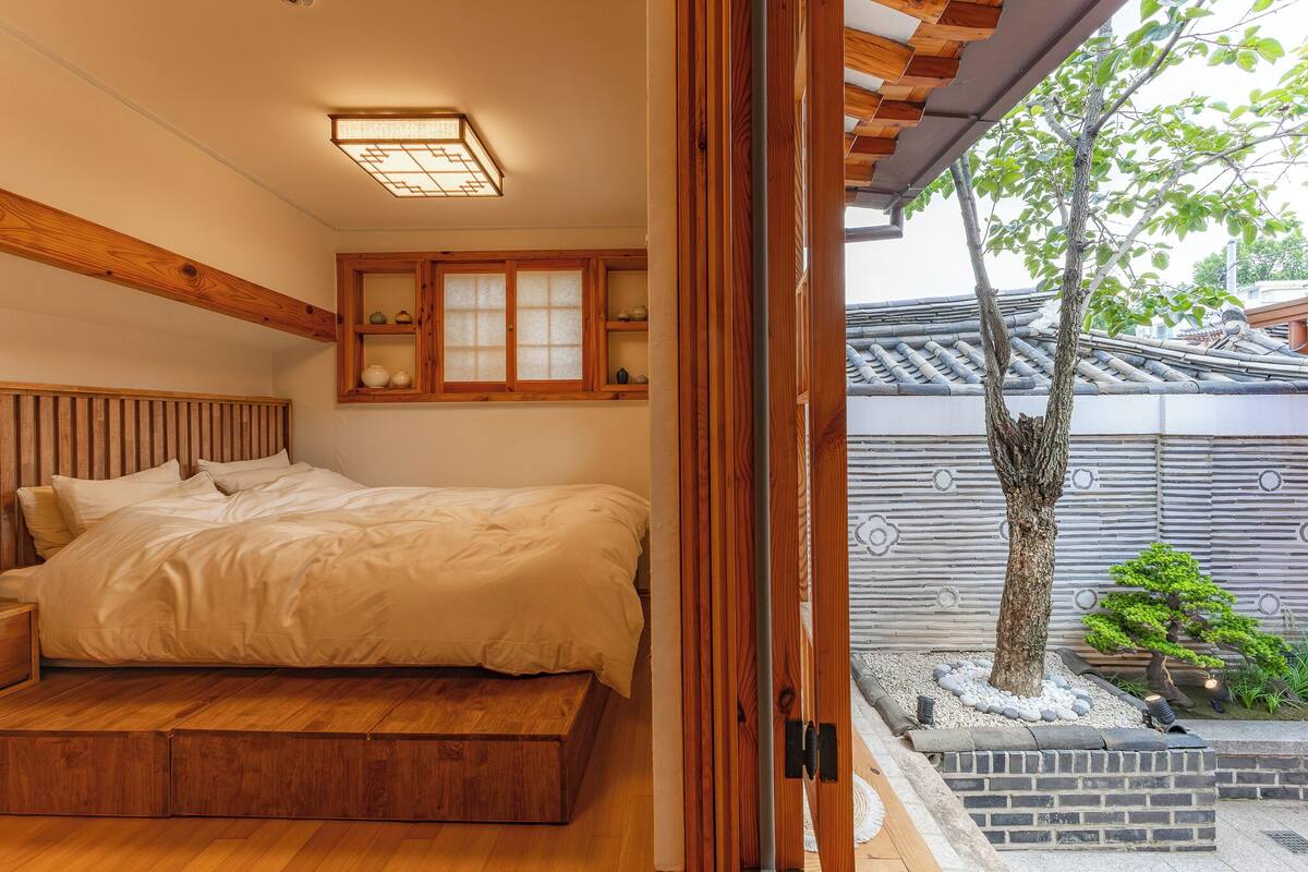 A bedroom with a wooden bed, a built-in shelf with decorative items, and a view of a traditional-style building and a tree outside.