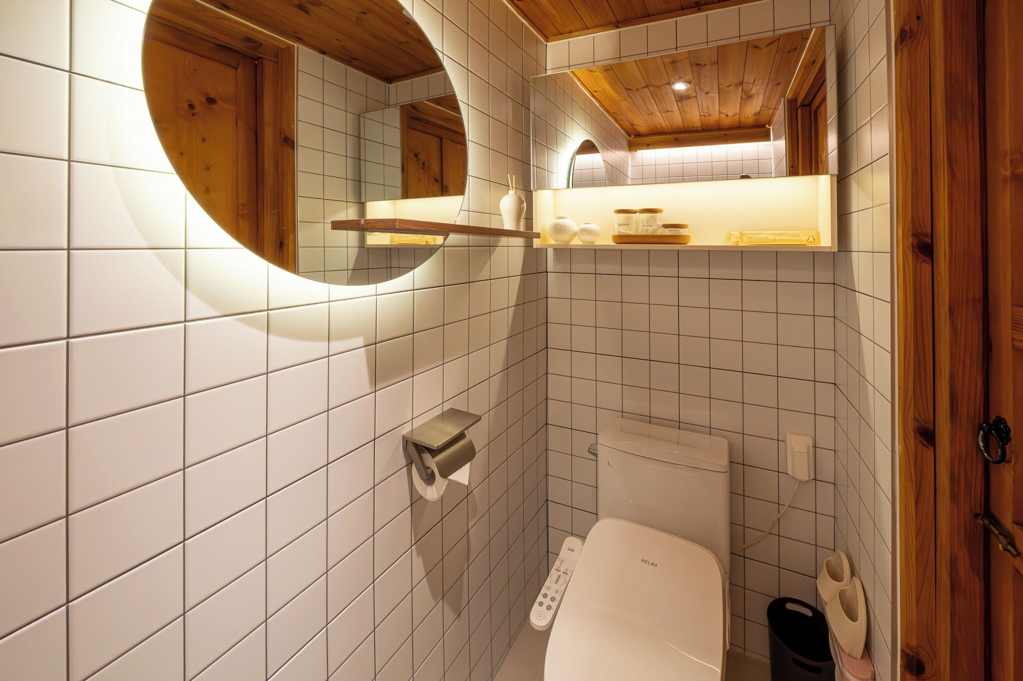 Een badkamer met witte betegelde wanden, een houten plafond, een toilet met bedieningspaneel, een spiegel en een plank met toiletartikelen.