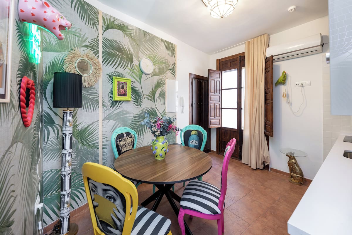 A dining area with a round wooden table, colorful chairs, a decorative wall with palm leaf patterns, a wall-mounted phone, and a small table with a glass top.