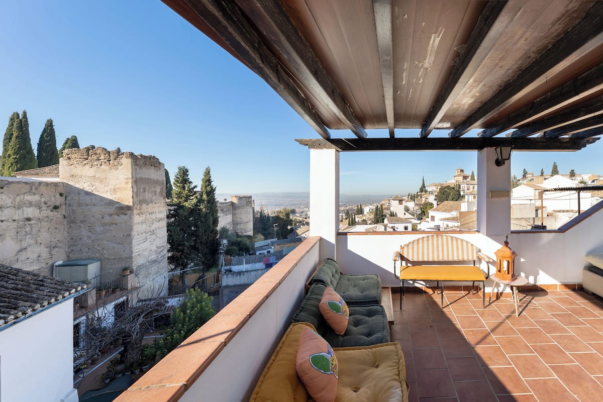 A rooftop terrace with a view of the city, a wooden pergola, and a seating area with cushions.