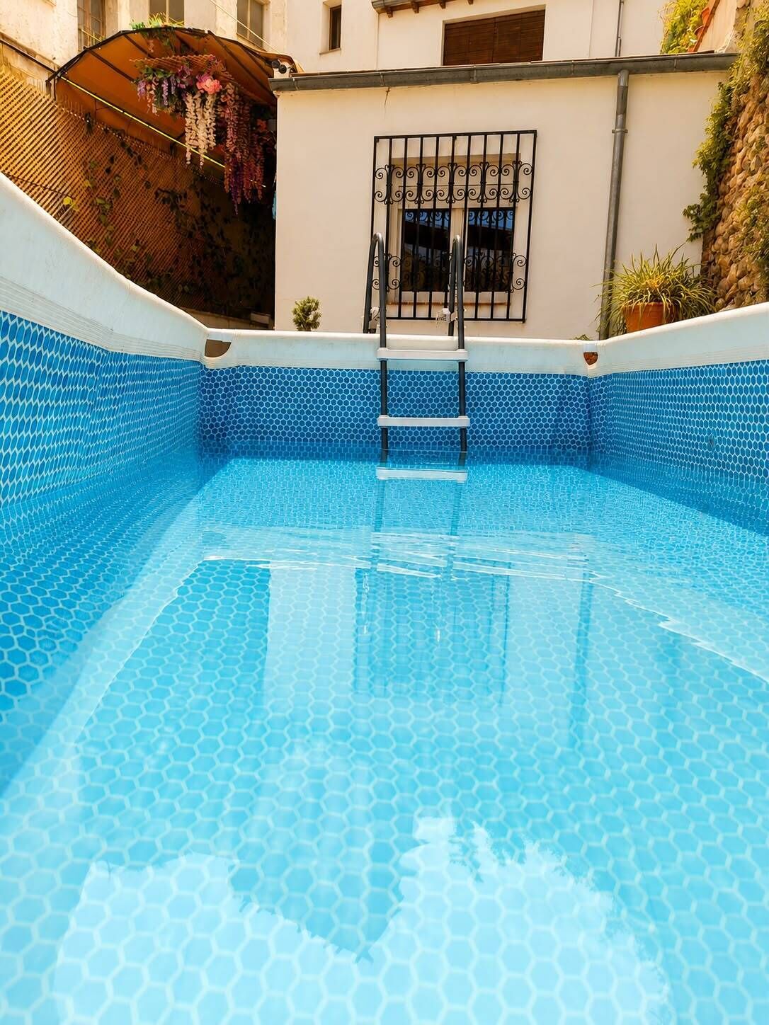 A clear blue swimming pool with a hexagonal tile pattern, a ladder leading into the pool, and a small window with decorative iron bars.