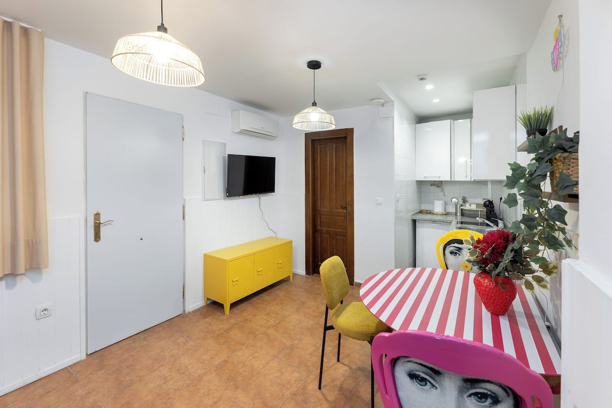 A compact kitchen with white cabinets, a yellow cabinet, and a dining area with a striped tablecloth and a red vase.