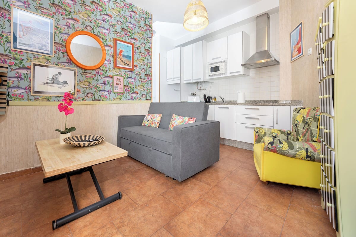 A compact living space with a grey sofa, a small wooden table with a bowl and plant, a yellow armchair, and a kitchen area with white cabinets and a microwave.