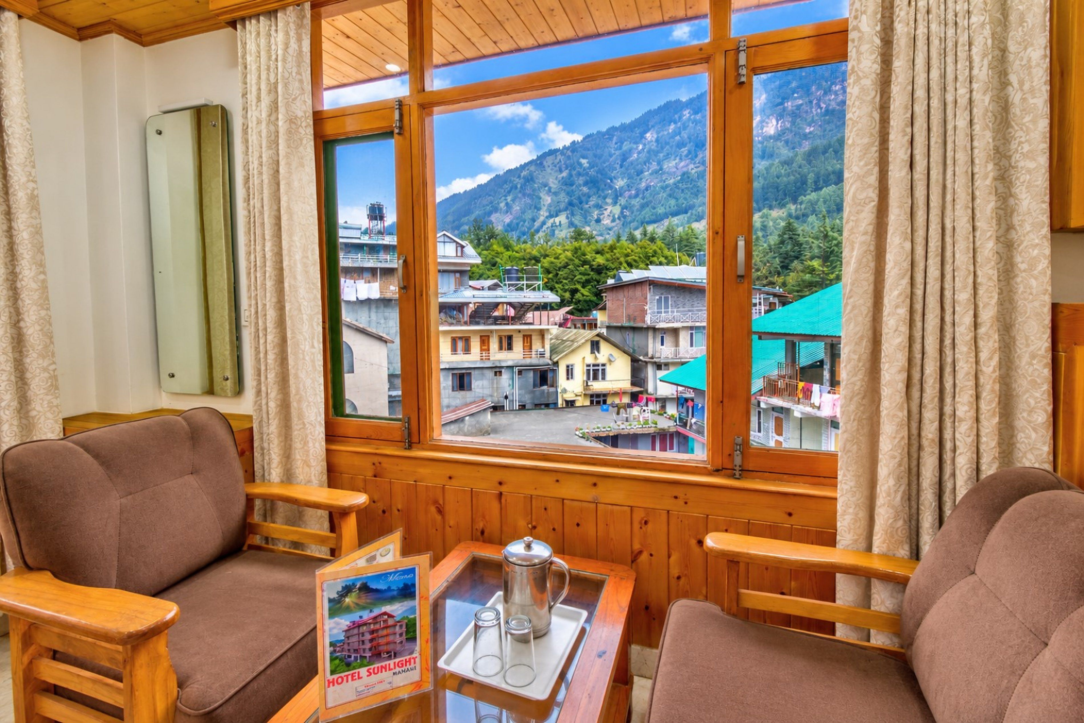 A room with a large window offering a view of a mountainous area and a village. There are two wooden chairs and a small table with a tray containing glasses and a container.