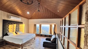 Un dormitorio con cama, un banco, una silla y ventanas grandes con cortinas.