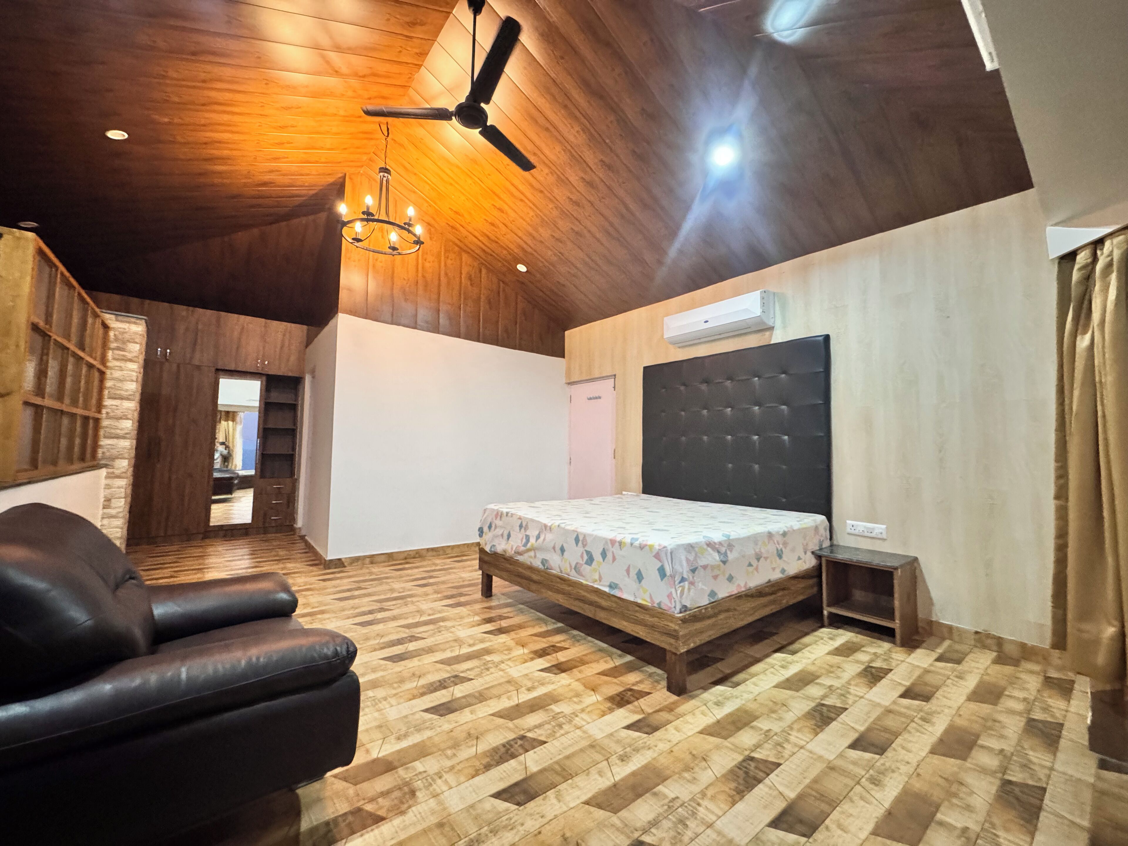 A bedroom with a wooden ceiling, a bed with a patterned bedspread, a black leather chair, and a ceiling fan.