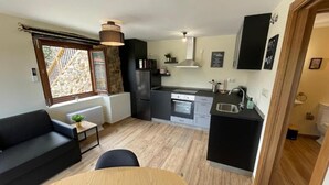 A compact kitchen with a dining area, a black sofa, and a window with a view of a stone building.