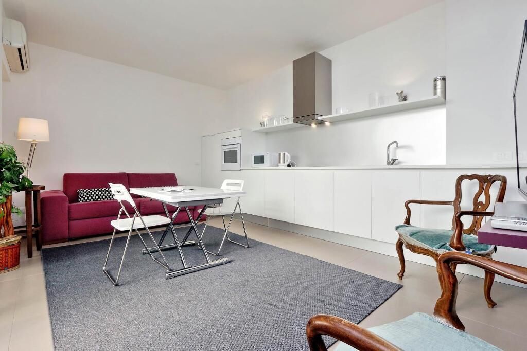 A modern living room with a red sofa, a dining table with chairs, a kitchen area with white cabinets, and a wooden chair.