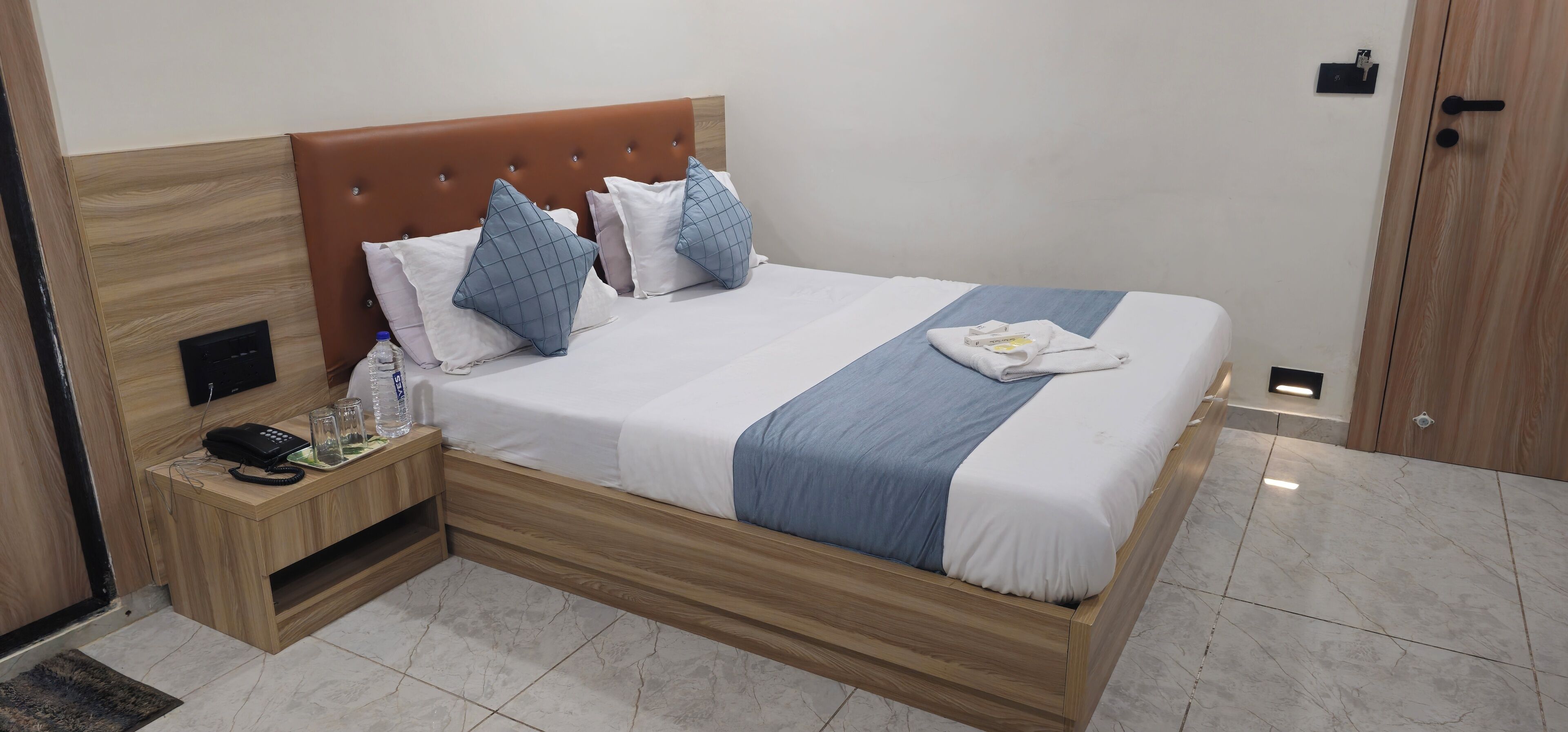 A neatly made bed with a wooden headboard, a bedside table with a phone and glasses, and a towel on the bed.