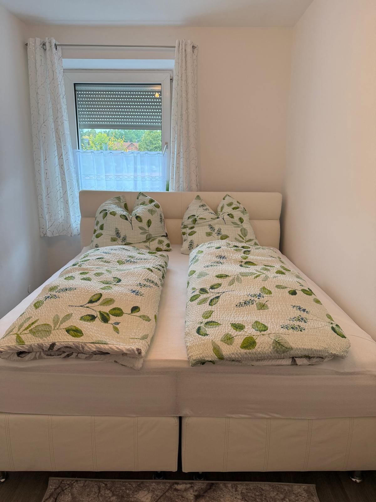 A double bed with a leaf-patterned duvet and pillows, a window with blinds, and a view of greenery outside.