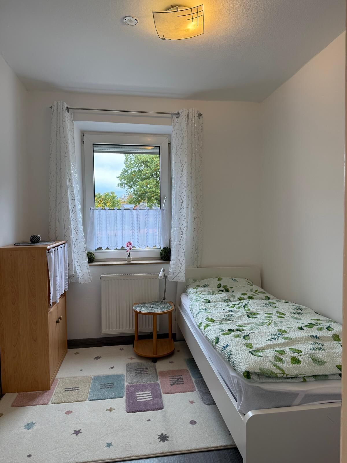 Ein kleines, ordentliches Schlafzimmer mit einem Einzelbett, einem Holzschrank, einem Heizkörper und einem Fenster mit Vorhängen.