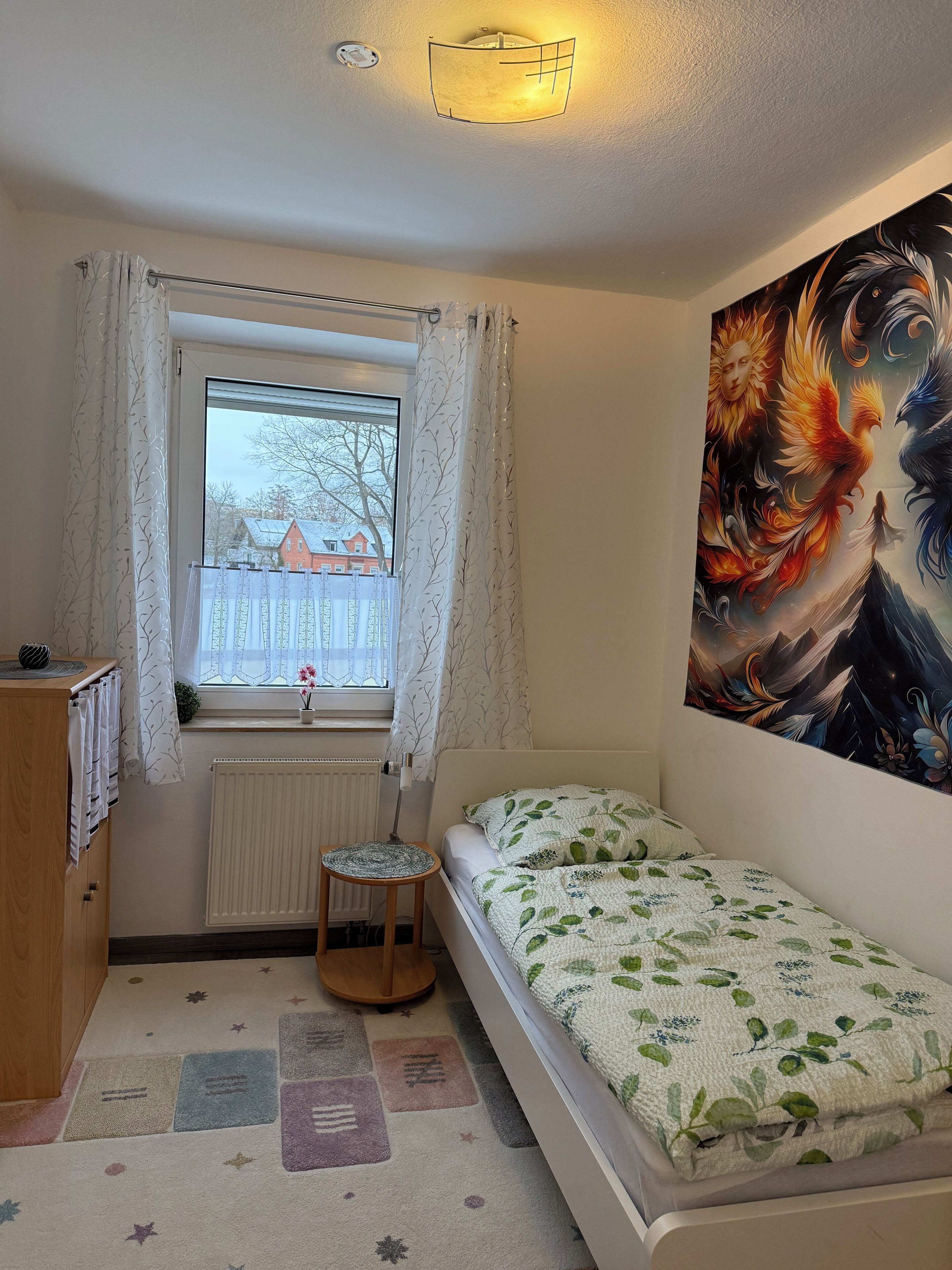Un dormitorio con cama, ventana con cortinas, radiador, un taburete pequeño, un cuadro colgado en la pared y un armario de madera.