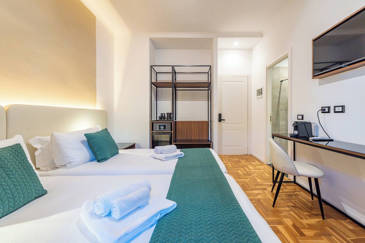 A hotel room with two beds, a desk, a chair, a mirror, and a kitchenette.
