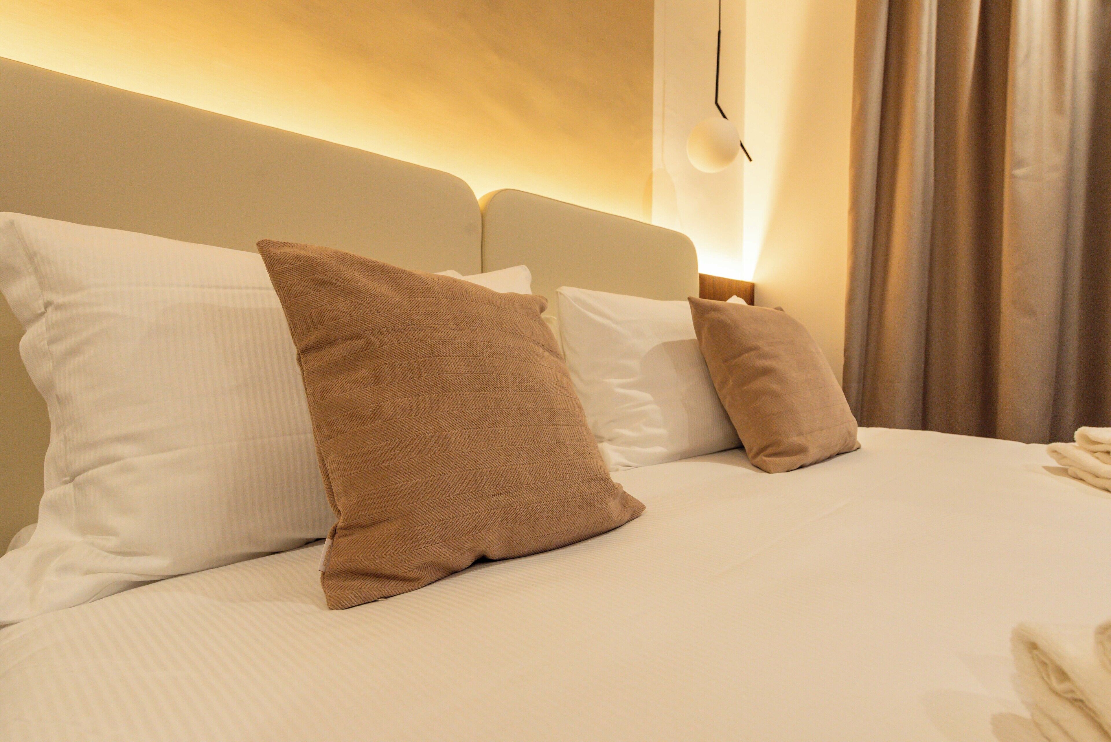 Una cama bien hecha con almohadas blancas y marrones, un cabecero y una lámpara fijada a la pared.