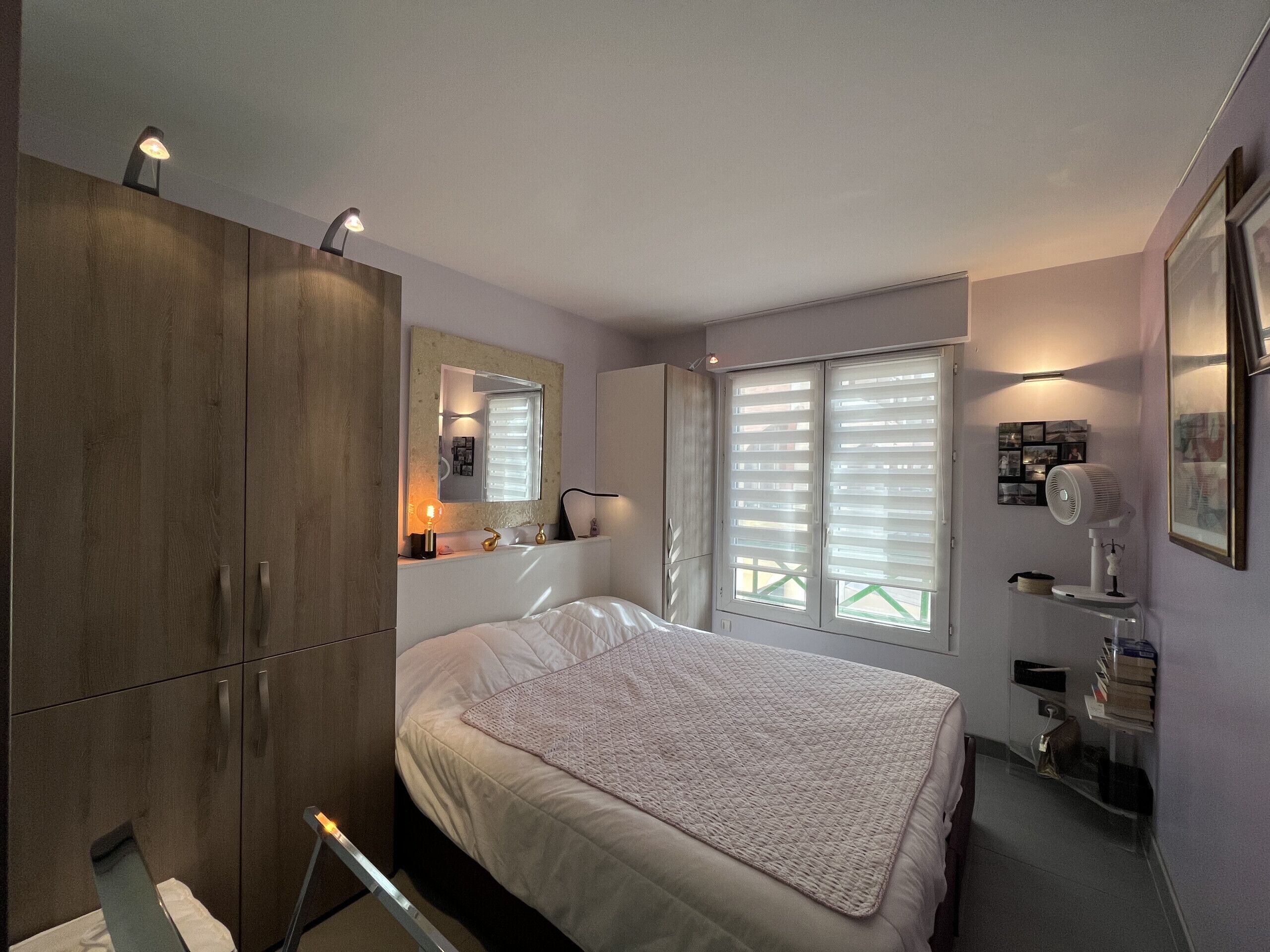 A bedroom with a bed, a wardrobe, a mirror, a window with blinds, and a bedside table with a lamp.