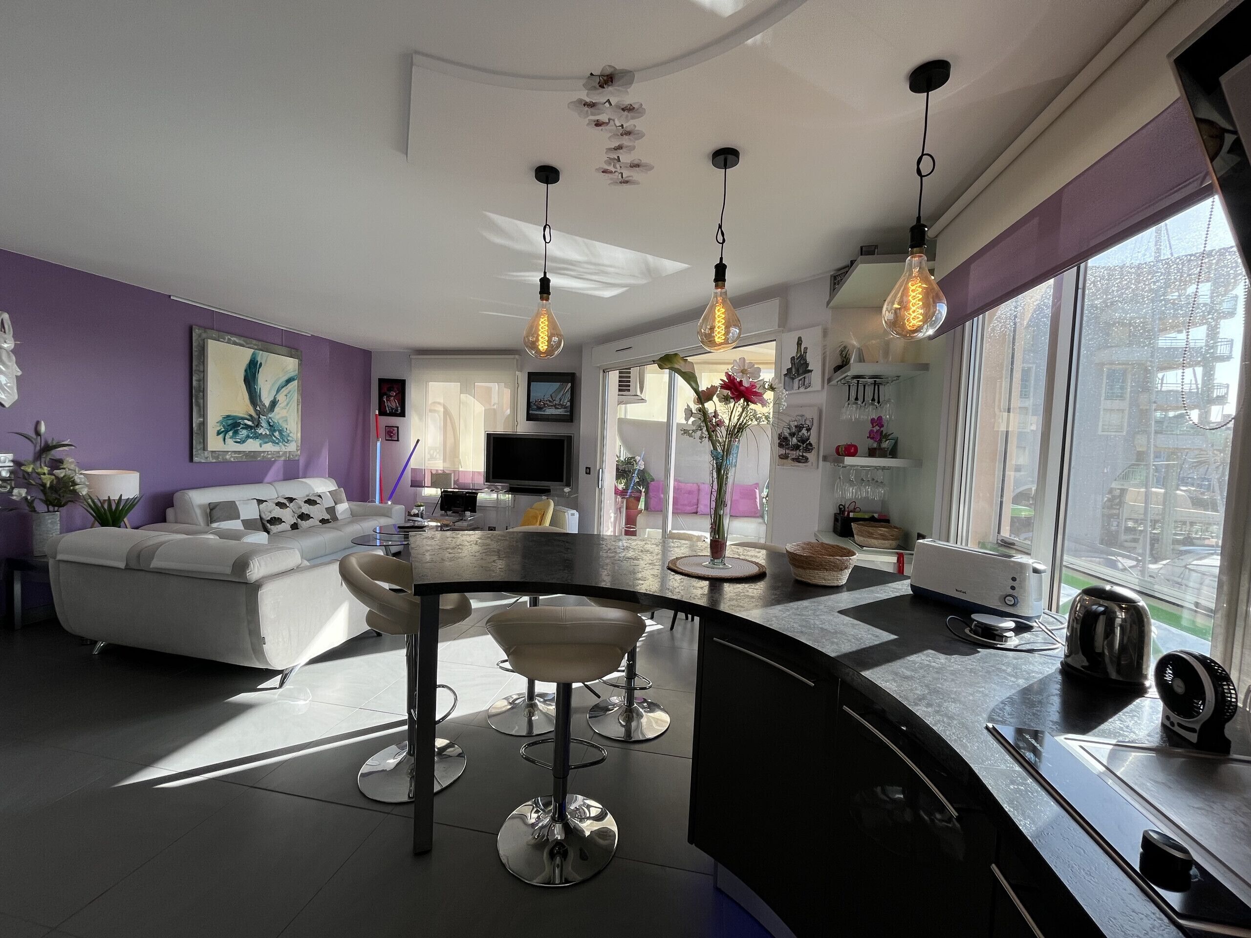 A modern living room with a sofa, a dining table, and a kitchen area with a kettle and a coffee machine.
