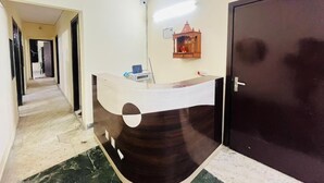 Un mostrador de recepción con acabado en madera oscura, una encimera blanca y un pequeño estante con un adorno. A la derecha hay una puerta cerrada, y un pasillo con suelo de baldosas se extiende en el fondo.