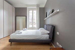 Ein modernes Schlafzimmer mit grauem Bett, weißen Bettwäsche, einem grauen Kopfteil, einem Fenster mit weißen Vorhängen und einem eingebauten Kleiderschrank.