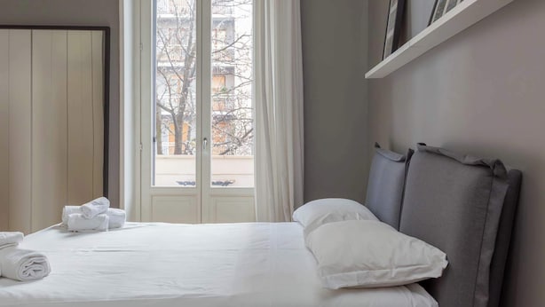 Ett hotellrum med en säng, ett fönster med gardiner, en sänggavel och en hylla med inramade bilder.