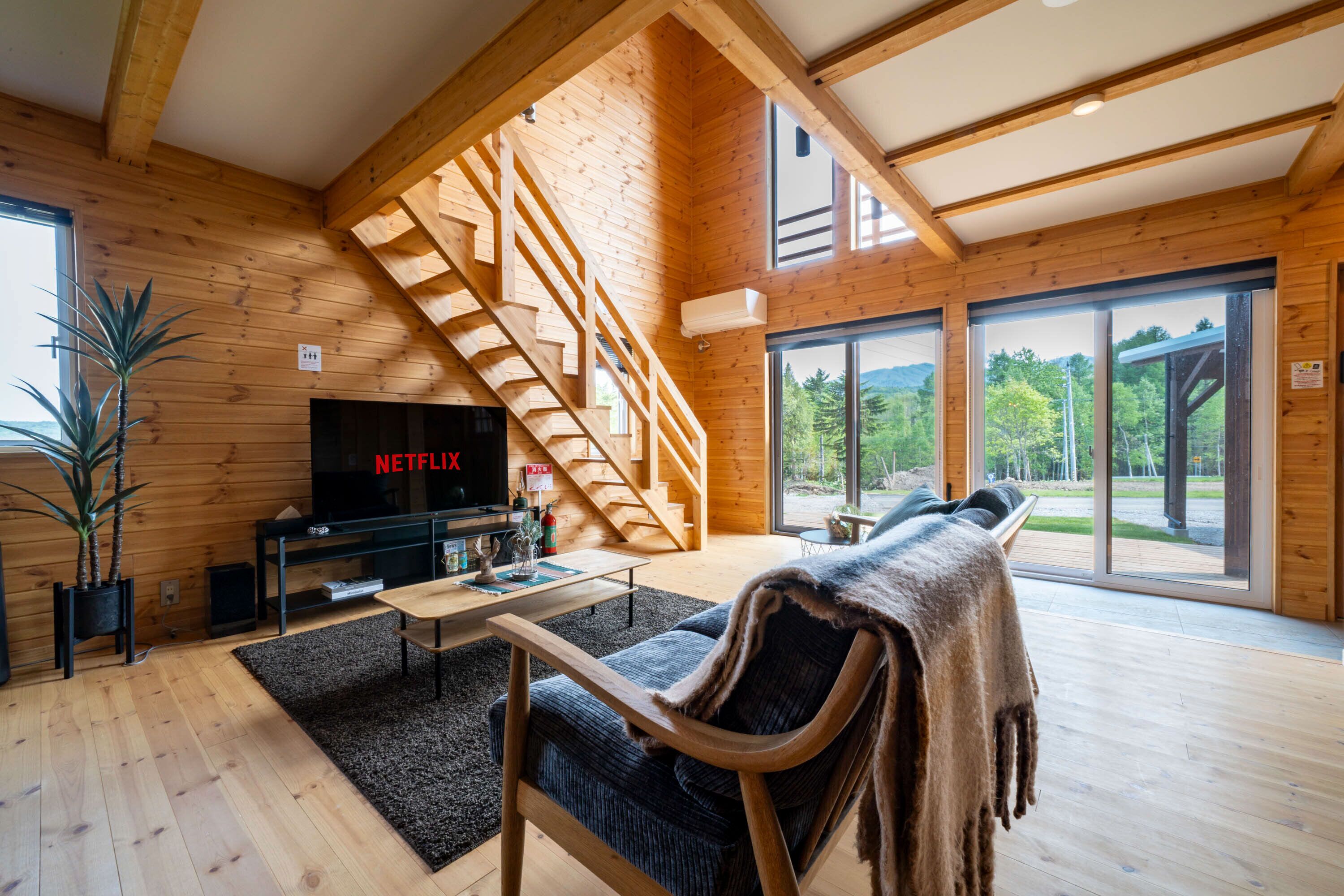 A modern log cabin interior with a wooden staircase, a flat-screen TV, a sofa, and large windows offering a view of the outdoors.