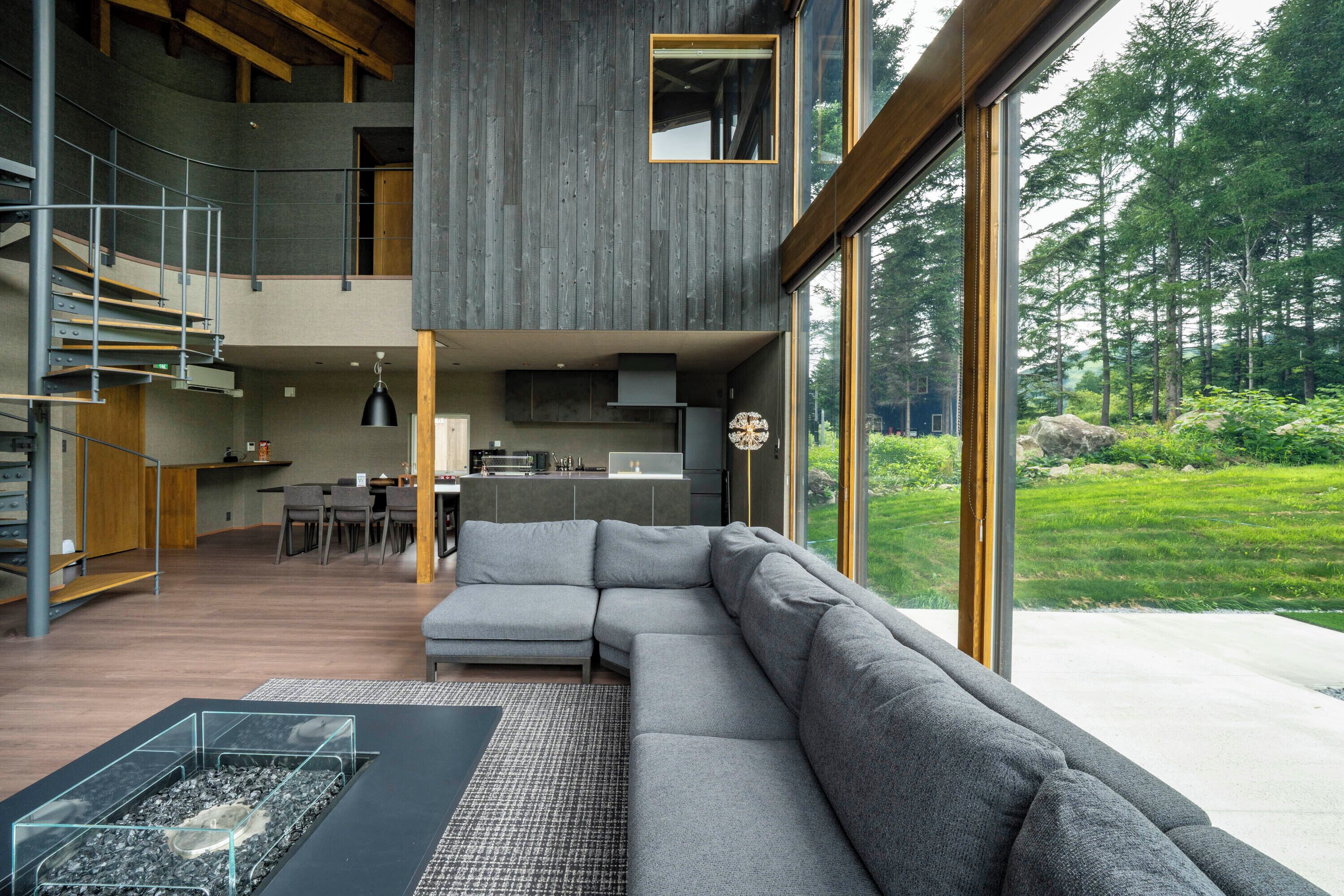 A modern interior with a grey sectional sofa, a glass coffee table, and a view of a forest through large windows.
