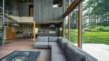 A modern interior with a grey sectional sofa, a glass coffee table, and a view of a forest through large windows.