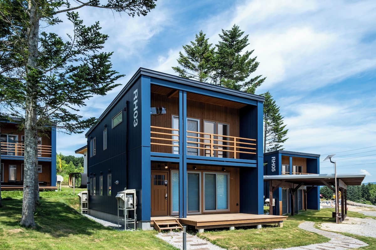 A modern two-story building with a blue exterior and wooden accents, featuring a balcony and a covered outdoor seating area.