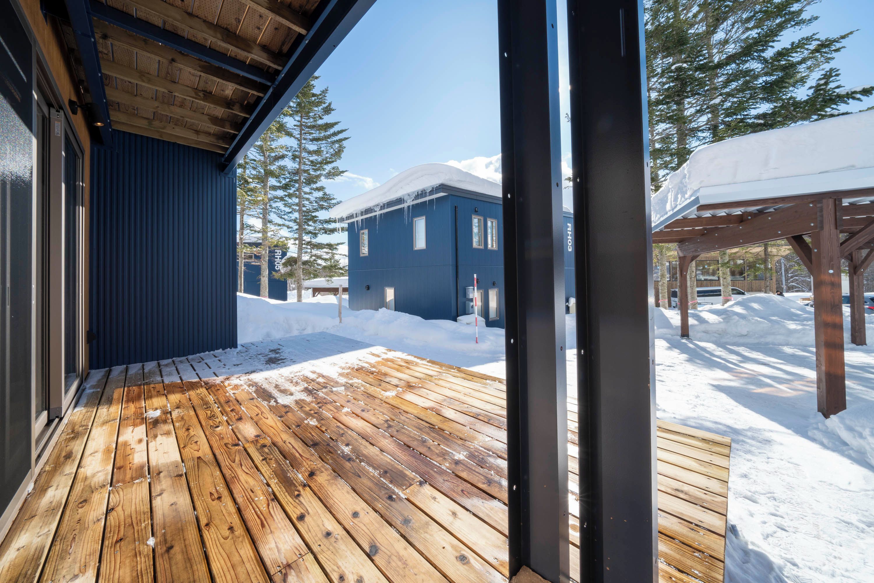 A wooden deck with snow on the ground, a blue building, and a covered area with snow.