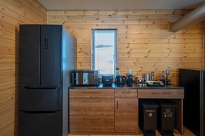 A compact kitchen with wooden walls, a black refrigerator, a microwave, a coffee maker, and waste bins labeled for recyclables and bottles.