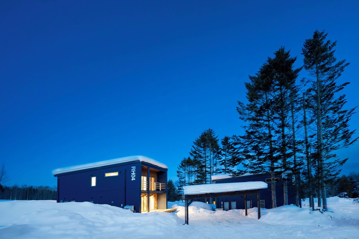 A modern building with a dark exterior and a lit entrance, surrounded by snow and tall trees.