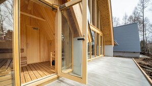 Una sauna de madera con puertas de vidrio, una terraza de madera y un edificio moderno al fondo.